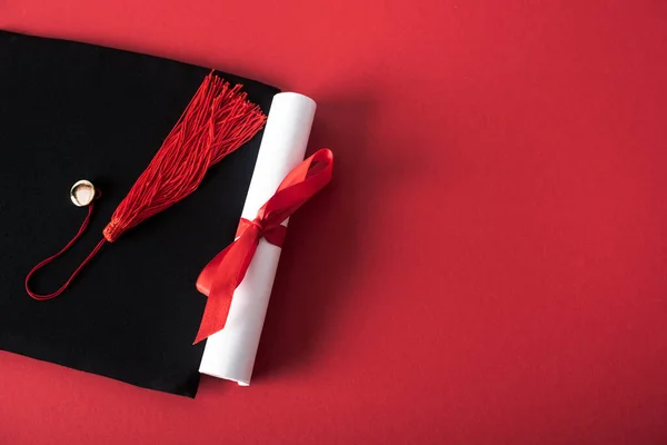 Top view of diploma with beautiful bow and graduation cap with tassel on red background — Stock Photo