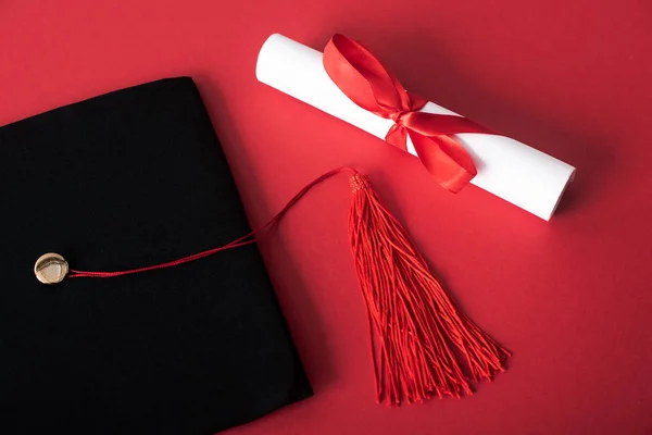 Top view of diploma with beautiful bow and black graduation cap with tassel on red background — Stock Photo