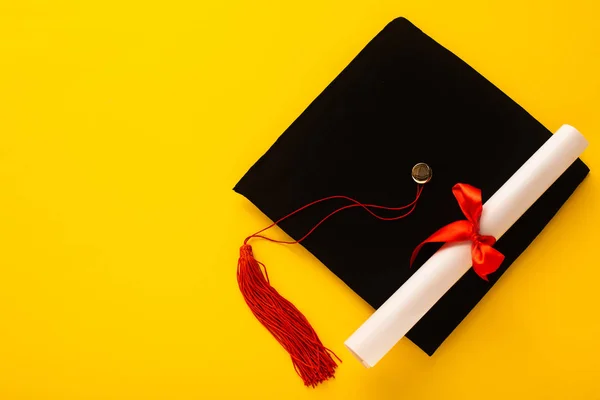 Vista superior de la gorra de graduación negra con borla roja con diploma en la parte superior sobre fondo amarillo - foto de stock