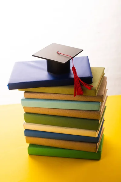 Graduation cap with red tassel on top of books on yellow surface on white background — Stock Photo