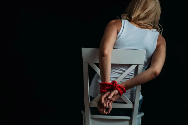 Back view of woman with tied hands sitting on white chair isolated on black — Stock Photo