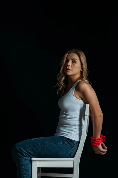 Side view of woman with tied hands looking at camera and sitting on white chair isolated on black — Stock Photo