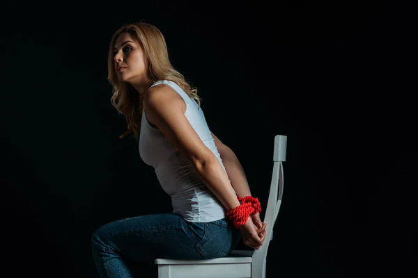 Side view of woman with tied hands looking away, sitting on chair isolated on black — Stock Photo