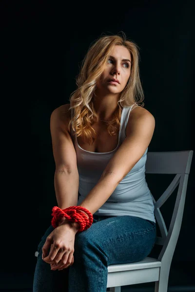 Femme les mains liées regardant ailleurs, assise sur une chaise isolée sur du noir — Photo de stock