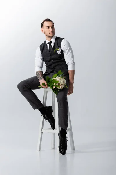 Handsome tattooed elegant bridegroom with bouquet sitting on highchair on grey — Stock Photo