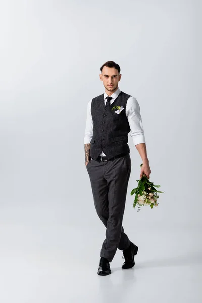 Handsome tattooed elegant bridegroom walking with bouquet on grey — Stock Photo