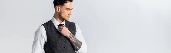 Handsome tattooed elegant bridegroom adjusting tie isolated on grey, panoramic shot — Stock Photo