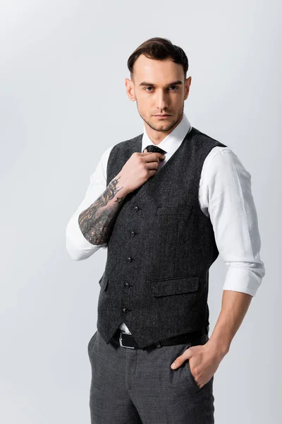 Handsome tattooed elegant bridegroom with hand in pocket adjusting tie isolated on grey — Stock Photo