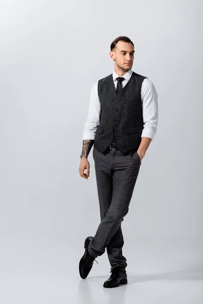 Guapo tatuado elegante novio posando con la mano en el bolsillo sobre fondo gris - foto de stock
