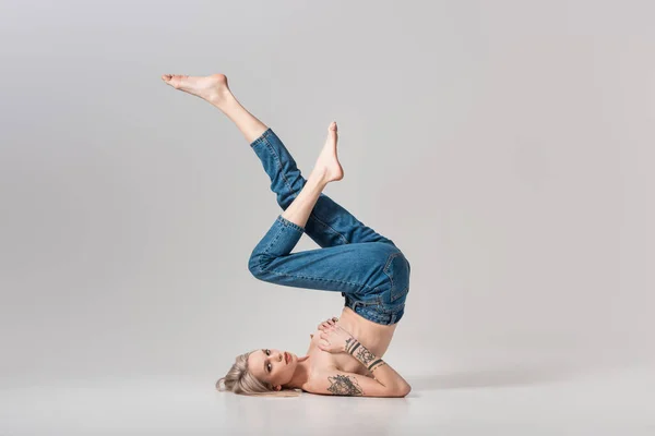 Joven chica tatuada en pantalones vaqueros posando en el suelo con las piernas levantadas en gris - foto de stock