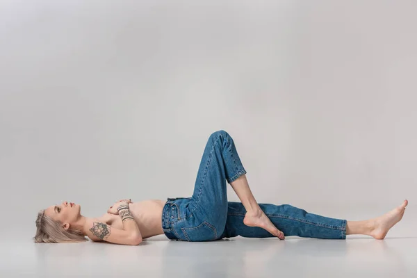 Joven topless tatuado chica en jeans acostado en el suelo en gris - foto de stock