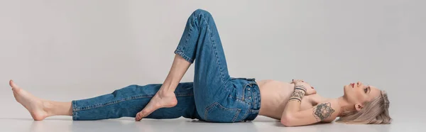 Young topless tattooed girl in jeans lying on floor on grey, panoramic shot — Stock Photo