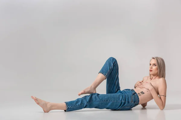 Young topless tattooed girl in jeans posing on floor on grey background — Stock Photo
