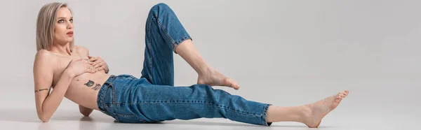 Jeune fille tatouée seins nus en jeans posant sur le sol sur fond gris, vue panoramique — Photo de stock