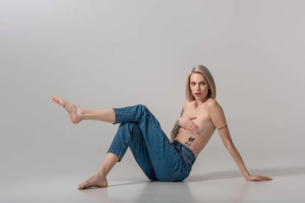 Young topless tattooed girl in jeans posing on floor on grey background — Stock Photo