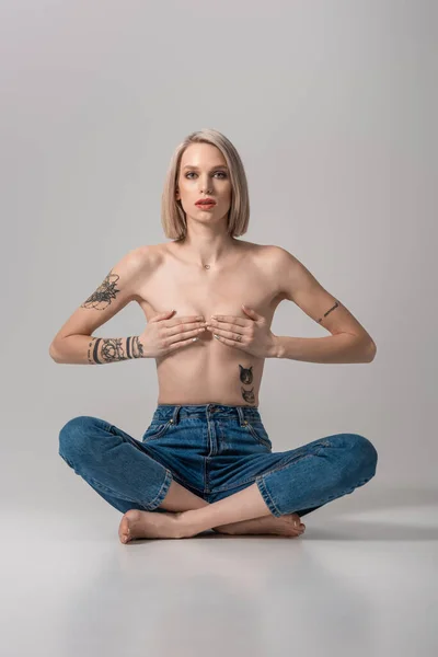 Sexy young topless tattooed woman covering breast with hands while sitting in lotus pose on grey — Stock Photo