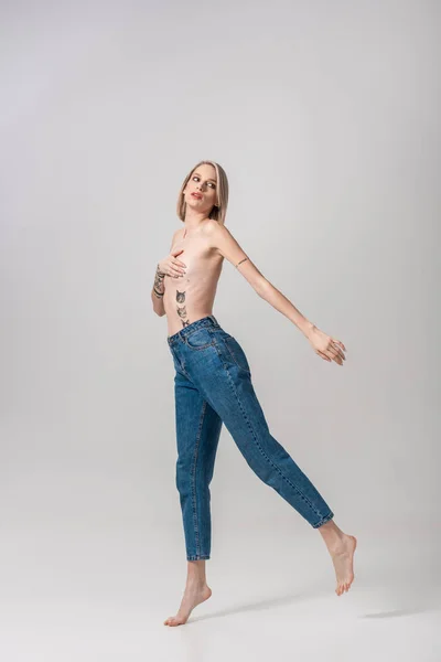 Sexy young topless tattooed woman covering breast with hand on tiptoe on grey — Stock Photo