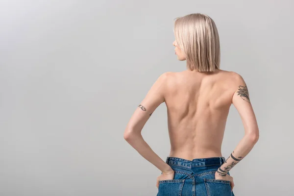 Vue arrière de sexy jeune femme tatouée seins nus posant avec les mains dans des poches isolées sur gris — Photo de stock