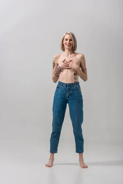 Feliz sexy joven en topless mujer tatuada que cubre el pecho con las manos en gris - foto de stock