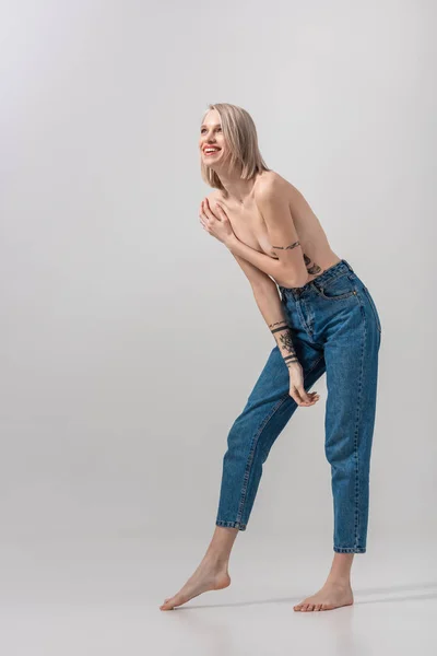 Happy sexy young topless tattooed woman covering breast with hand on grey — Stock Photo