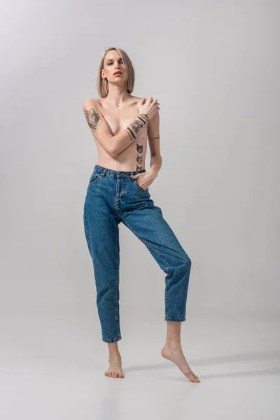 Sexy joven en topless mujer tatuada que cubre el pecho con la mano en gris - foto de stock