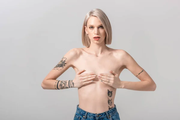 Sexy joven en topless mujer tatuada que cubre el pecho con las manos aisladas en gris - foto de stock