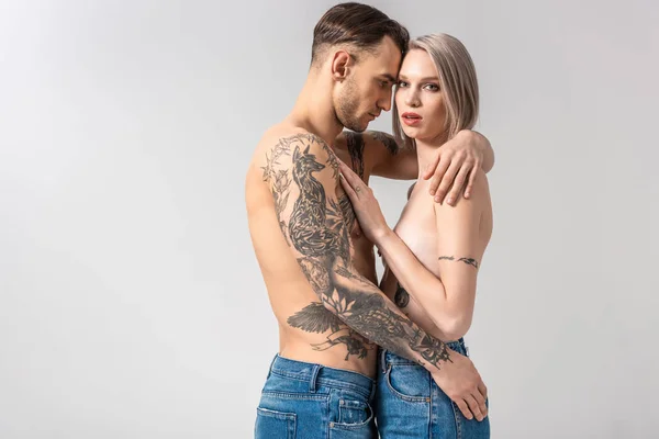 Вид сбоку на молодую обнаженную татуированную пару, обнимающую изолированную на сером — стоковое фото