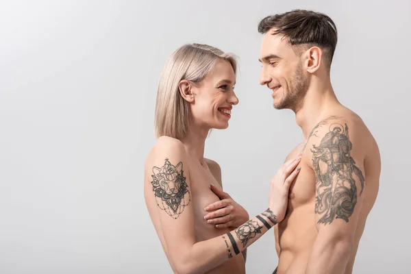 Vista lateral de feliz joven desnuda pareja tatuada abrazo aislado en gris - foto de stock