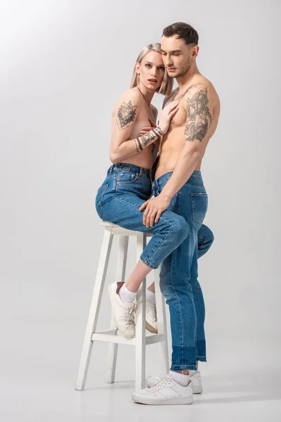 Joven pareja tatuada sin camisa en jeans abrazándose en silla en gris - foto de stock
