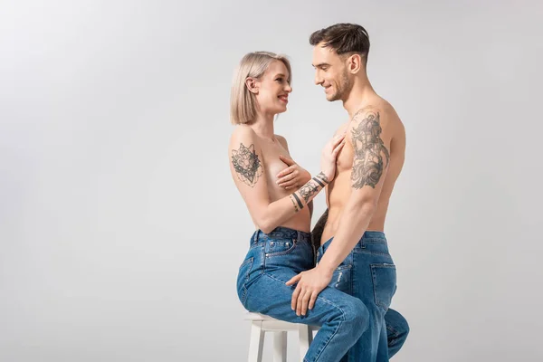 Vista laterale di felice giovane coppia tatuata senza maglietta in jeans che abbraccia sulla sedia isolata sul grigio — Foto stock