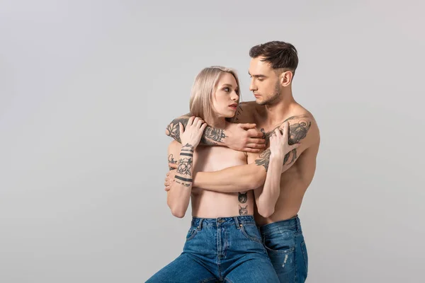 Jovem sem camisa tatuado casal em jeans abraçando isolado no cinza — Fotografia de Stock