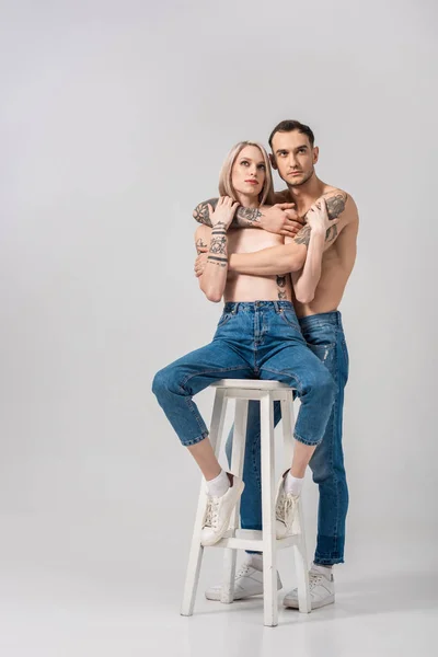 Junges, hemdloses tätowiertes Paar in Jeans umarmt sich auf Stuhl auf grau — Stockfoto