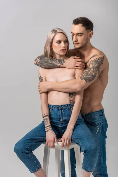 Junges tätowiertes Paar in Jeans umarmt sich auf Stuhl isoliert auf grau — Stockfoto