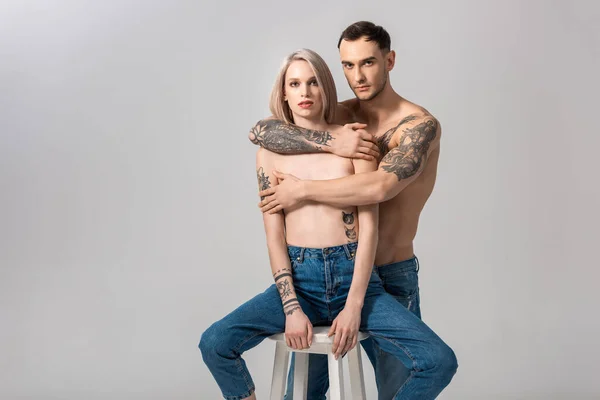 Молода татуйована пара без сорочки в джинсах обіймається на стільці ізольовано на сірому — стокове фото