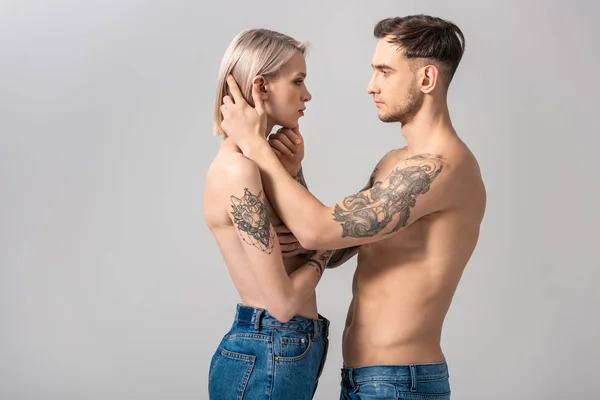Боковой вид молодой пары без рубашки с татуировками в джинсах, стоящей лицом к лицу, изолированной на сером — стоковое фото