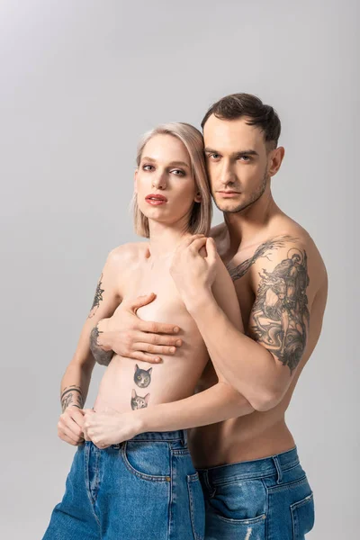 Молода татуйована пара без сорочки в джинсах обіймається ізольовано на сірому — стокове фото