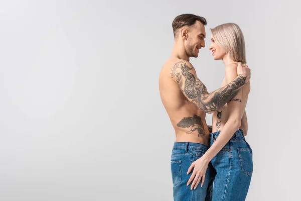 Вид збоку щасливої молодої сорочки татуювання пара в джинсах обіймається ізольовано на сірому — стокове фото