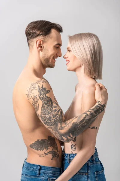 Side view of happy young shirtless tattooed couple in jeans standing face to face isolated on grey — Stock Photo