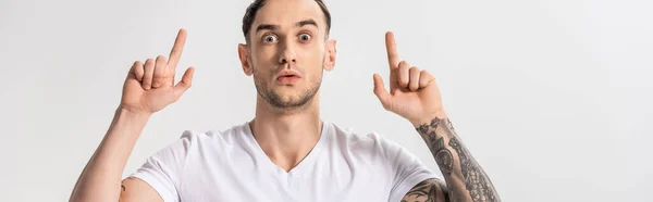 Потрясенный красивый молодой татуированный мужчина, указывающий пальцами на белый, панорамный снимок — Stock Photo