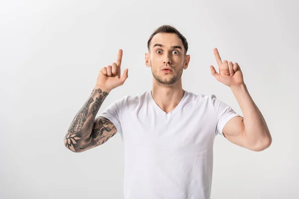 Sorprendido guapo joven tatuado hombre señalando con los dedos hacia arriba aislado en blanco - foto de stock
