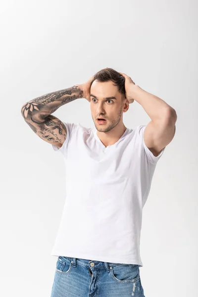Шокований красивий молодий татуйований чоловік з руками на голові ізольований на білому — стокове фото