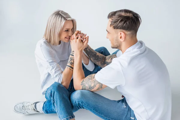 Happy young tattooed pregnant woman sitting with husband on floor and holding hands on grey — Stock Photo