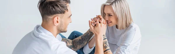 Lächelnde junge tätowierte Schwangere, die mit ihrem Mann auf dem Boden sitzt und die Hände isoliert auf grauen, panoramischen Aufnahmen hält — Stockfoto