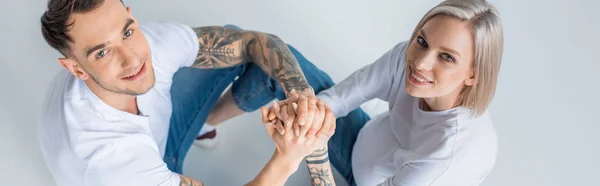 Vista aerea della giovane donna incinta tatuata sorridente seduta con il marito sul pavimento e che si tiene per mano sul grigio, scatto panoramico — Foto stock