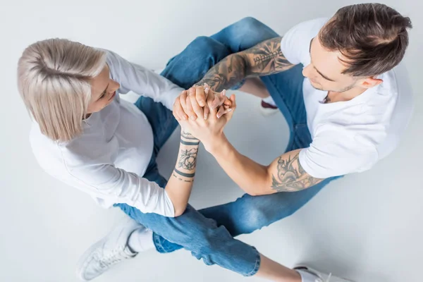 Overhead view of young tattooed pregnant woman sitting with husband on floor and holding hands on grey — Stock Photo