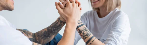 Corte vista de sorrir jovem tatuado mulher sentado com o marido no chão e de mãos dadas isolado em cinza, tiro panorâmico — Fotografia de Stock