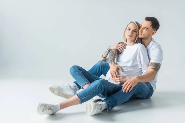 Молода татуйована вагітна жінка сидить з чоловіком на підлозі на сірому — стокове фото