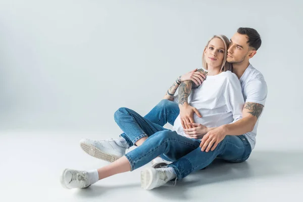 Young tattooed pregnant woman sitting with husband on floor on grey — Stock Photo