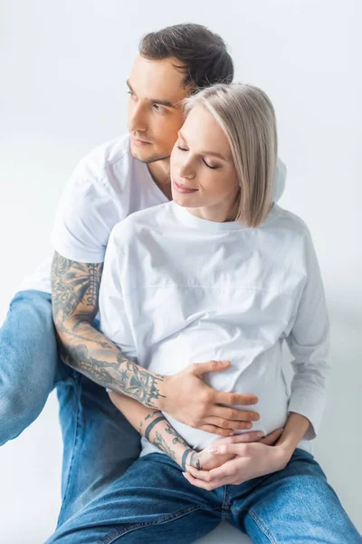 Jovem mulher grávida tatuada sentado com o marido no chão em cinza — Fotografia de Stock