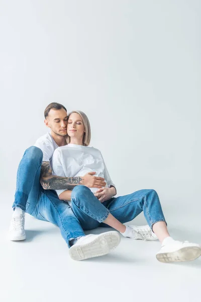 Молода татуйована вагітна жінка з закритими очима, сидячи з чоловіком на підлозі на сірому — стокове фото